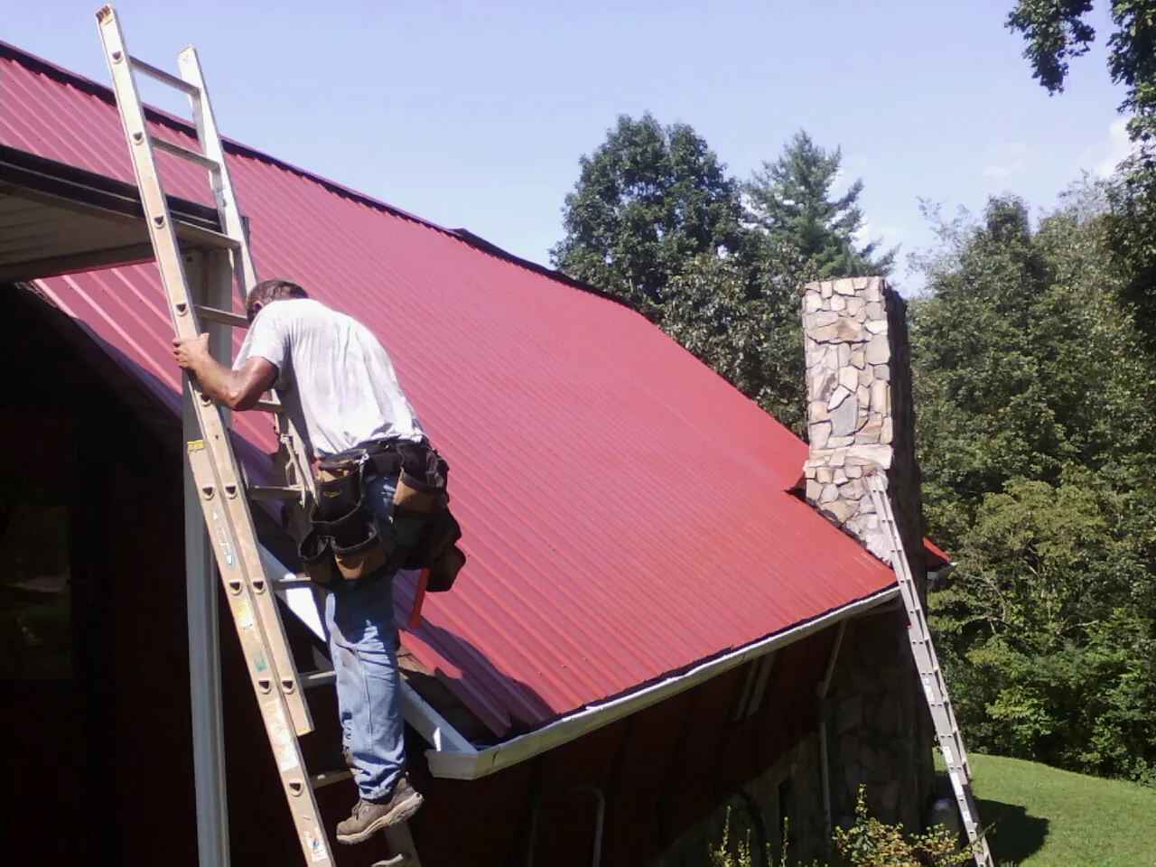 Roofer on a ladder beside a red metal roof and stone chimney — Soffit & Fascia Repair by Roofing Brothers in Agency